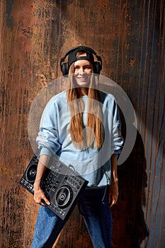 A young smiling long-haired DJ girl in a blue sweater, jeans, a black baseball cap and black headphones stands against a