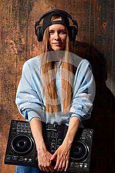 A young smiling long-haired DJ girl in a blue sweater, jeans, a black baseball cap and black headphones stands against a