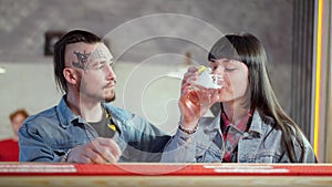 Young smiling loving man with tattoo on face and piercing admiring beautiful relaxed woman drinking alcohol drink in pub