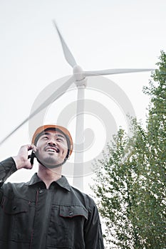 Young smiling male engineer in a hardhat on the phone beside a wind turbine