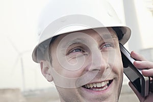 Young smiling male engineer in a hardhat on the phone beside a wind turbine, close-up