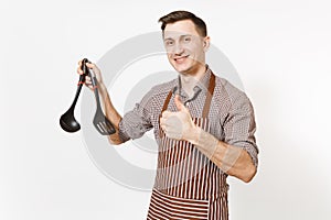 Young smiling man chef or waiter in striped brown apron, shirt holding black ladle or kitchen spoon, spatula isolated on