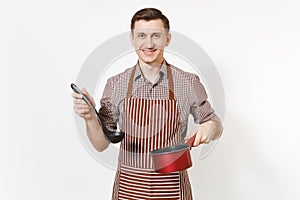 Young smiling man chef or waiter in striped brown apron, shirt holding red empty stewpan black ladle isolated on white