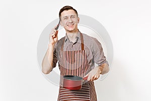 Young smiling man chef or waiter in striped brown apron, shirt holding red empty stewpan black ladle isolated on white