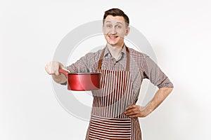 Young smiling man chef or waiter in striped brown apron, shirt holding red empty stewpan, pan or pot isolated on white