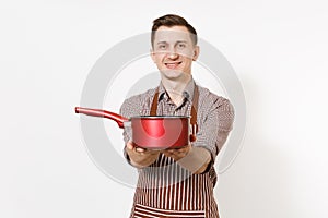 Young smiling man chef or waiter in striped brown apron, shirt holding red empty stewpan, pan or pot isolated on white