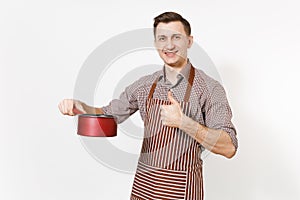 Young smiling man chef or waiter in striped brown apron, shirt holding red empty stewpan, pan or pot isolated on white