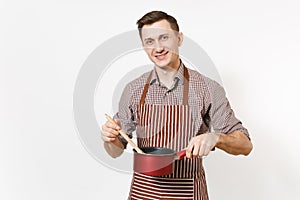 Young smiling man chef or waiter in striped brown apron, shirt holding red empty stewpan, wooden spoon isolated on white