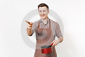 Young smiling man chef or waiter in striped brown apron, shirt holding red empty stewpan, wooden spoon isolated on white