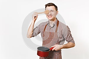 Young smiling man chef or waiter in striped brown apron, shirt holding red empty stewpan, wooden spoon isolated on white