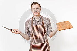 Young smiling man chef or waiter in striped brown apron, shirt holding wooden cutting board, knife isolated on white