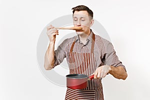 Young smiling man chef or waiter in striped brown apron, shirt tasting red empty stewpan, wooden spoon isolated on white
