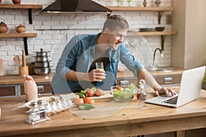 Young smiling man cooking breakfast in the kitchen checking recipe in his laptop , sunday morning