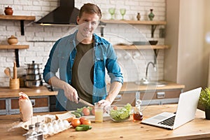 Young smiling man cutting tomatoes with knife while cooking breakfast in the kitchen checking recipe in his laptop , sunday