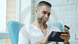 Young smiling man doing online shopping using digital tablet computer sitting at balcony at home