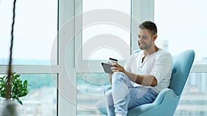 Young smiling man doing online shopping using digital tablet computer sitting at balcony in modern loft apartment