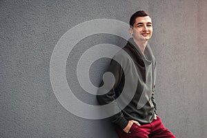 Young smiling man looking at camera against grey wall. Guy wearing casual clothing outdoors.