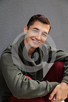 Young smiling man looking at camera against grey wall. Guy wearing casual clothing outdoors.