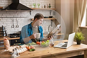 Young smiling man reading magazine while having breakfast in well-equipped kitchen checking recipe in his laptop , sunday morning