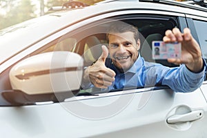 Young smiling man sitting in the car and showing his new driver license with thumb up sign