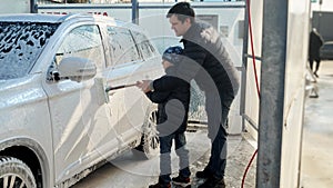 Young smiling man with son washing car outdoors with brush and foam. Automobile care, transport cleaning, dirty car.