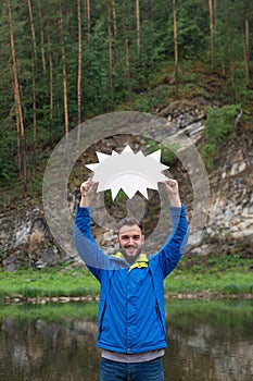 Young smiling man standing on bank of river keeping blank white board in hands above his head, rocky cliffs on background