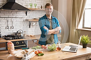 Young smiling man standing in well-equipped kitchen while having breakfast checking recipe in his laptop , sunday morning