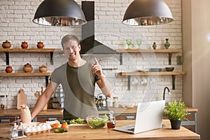 Young smiling man standing in well-equipped kitchen looking for interesting ideas for breakfast checking recipe in his laptop ,