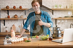 Young smiling man standing in well-equipped kitchen looking for interesting ideas for breakfast checking recipe in his laptop