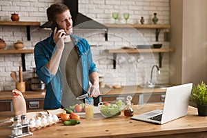 Young smiling man talking on the phone while cooking breakfast in the kitchen checking recipe in his laptop , sunday morning
