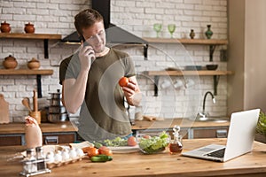 Young smiling man talking on the phone with tomatoe in his habd while cooking breakfast in the kitchen checking recipe in his