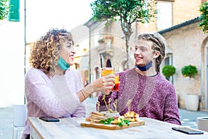 Young smiling millenial friends toasting a cocktail in a bar restaurant outdoor wearing protective face mask during happy hour in