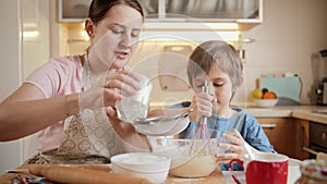 Young smiling mother cooking with little son and sifting flour with sieve. Children cooking with parents, little chef