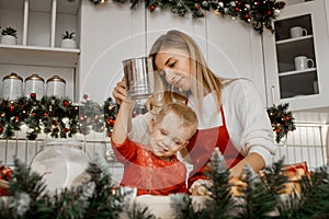 Young smiling mother cooking with little son and sifting flour with sieve in the christmas decorated kitchen