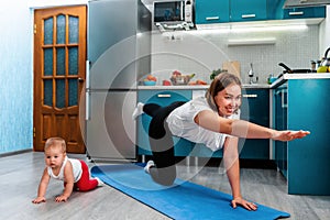 A young smiling mother does a yoga asana on a mat while her baby crawls next to her on the floor. Sports and yoga at home with