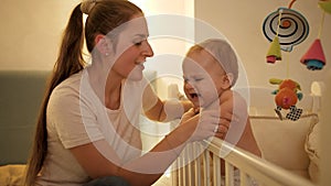 Young smiling mother entertaining crying baby son shouting in crib at night. Concept of parenting, children development.