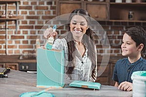 Young smiling mother helping her little son paint a wooden birdhouse