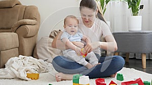 Young smiling mother hugging and holding her baby son playing with toy cars on floor in living room. Concept of family
