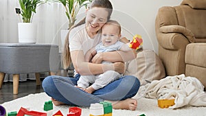 Young smiling mother hugging and holding her baby son playing with toy cars on floor in living room. Concept of family