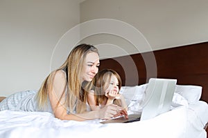 Young smiling mother lying in bed with little daughter and using laptop.