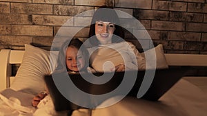 Young smiling mother lying in bed with little daughter and using laptop.