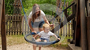 Young smiling mother swinging her cute toddler boy in rope nest swing at park playground