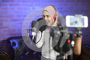 Young smiling muslim female singer wearing headphones with a microphone while recording song in a music studio with colorful