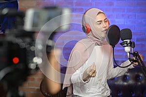 Young smiling muslim female singer wearing headphones with a microphone while recording song in a music studio with colorful