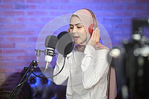 Young smiling muslim female singer wearing headphones with a microphone while recording song in a music studio with colorful