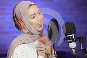 Young smiling muslim female singer wearing headphones with a microphone while recording song in a music studio with colorful