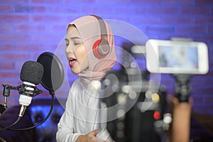 Young smiling muslim female singer wearing headphones with a microphone while recording song in a music studio with colorful