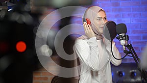 Young smiling muslim female singer wearing headphones with a microphone while recording song in a music studio with colorful