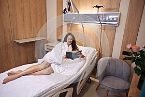 Young smiling pregnant woman reading book for future baby on medical couch