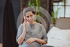 Young smiling pregnant woman touching belly talking on mobile phone with friend while spending time at home